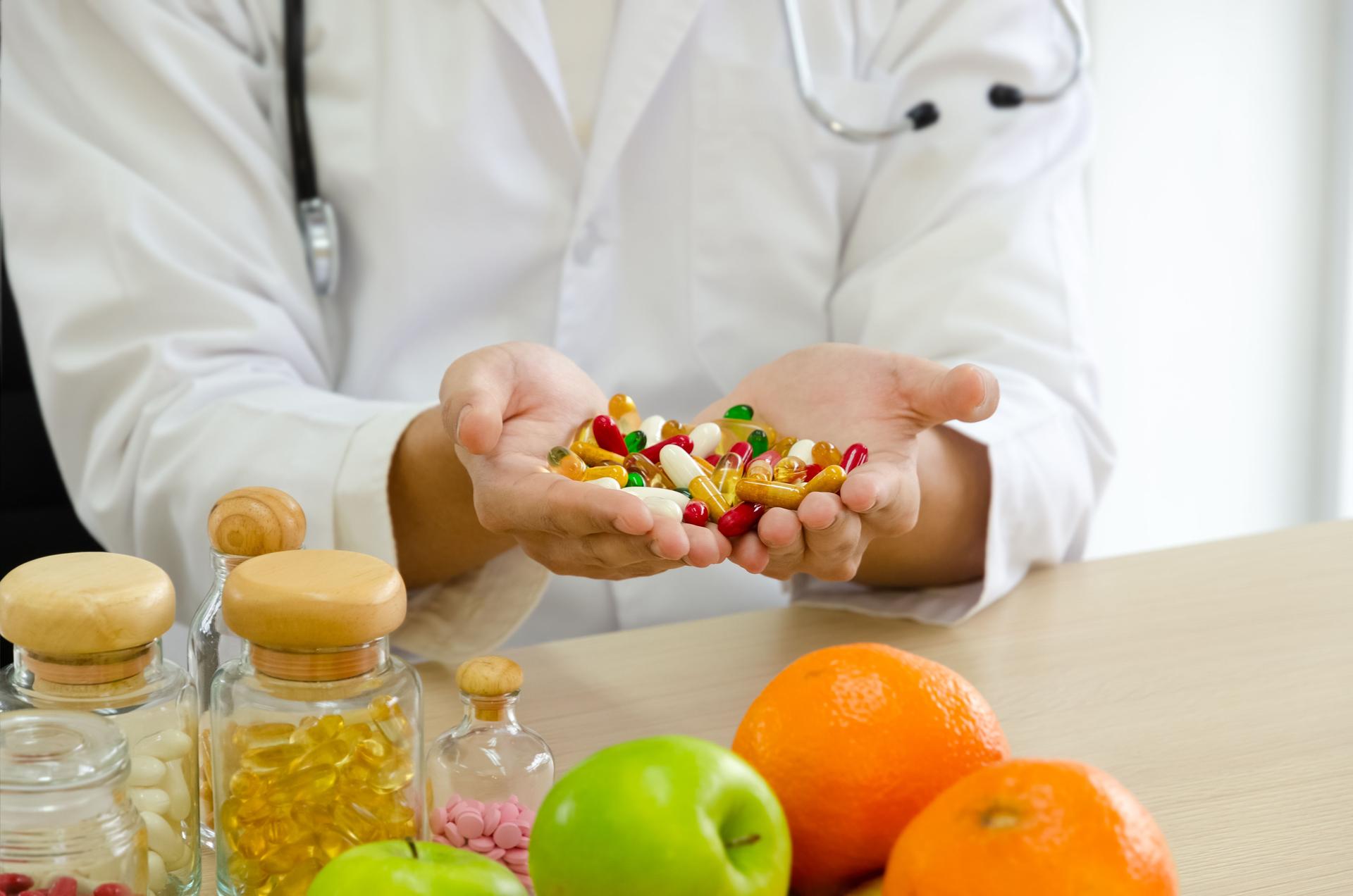 Nutritionist doctor in white coat with stethoscope holding colorful pills in open palms.  Jars of pills, apple and orange on clinic desk